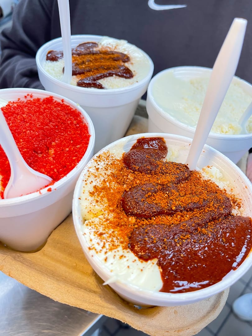 Four cups of esquites, two with chili powder and sauce, one with red sprinkles, and one plain.