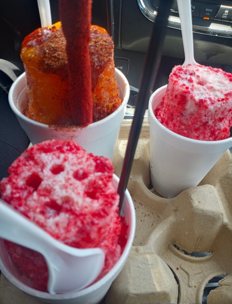 Three cups of colorful shaved ice, one mango with chili and two red with white toppings, in a car cup holder.